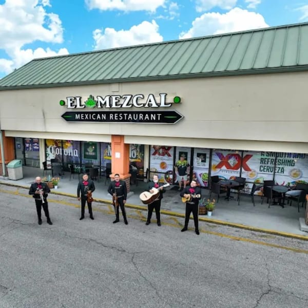Five mariachi band members stand outside El Mezcal Mexican Restaurant, holding instruments.