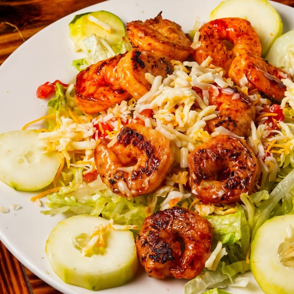 Grilled shrimp salad with shredded cheese, lettuce, and cucumber slices on a white plate.