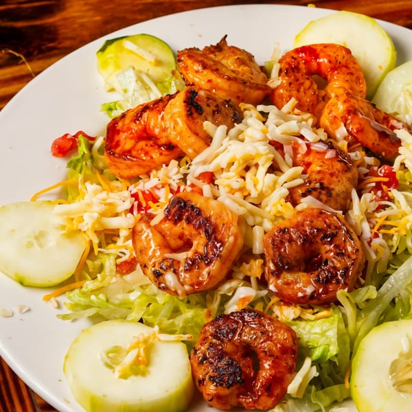 Grilled shrimp salad with shredded cheese, lettuce, and cucumber slices on a white plate.