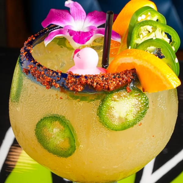 A large El Mezcal margarita with a chili-lime rim, orange slices, jalapeños, a pink rubber duck, and an orchid.