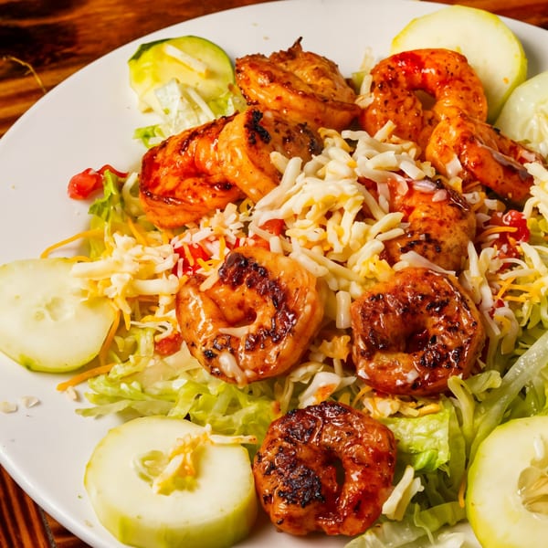 Grilled shrimp salad with shredded cheese, lettuce, and cucumber slices on a white plate.
