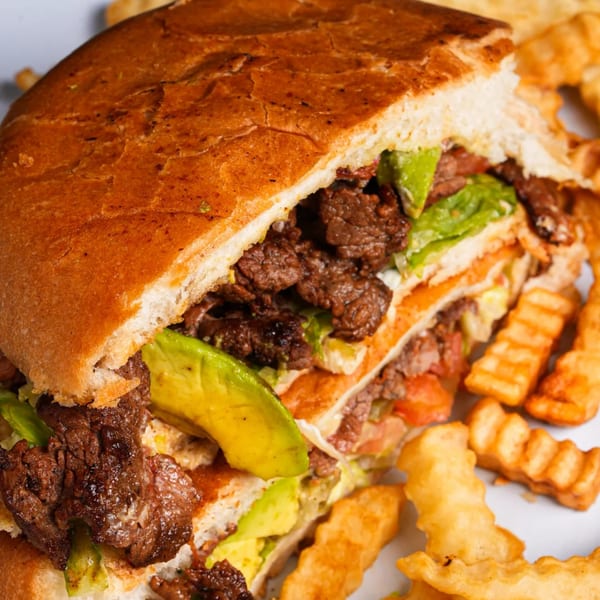 A large Mexican torta sandwich with steak, avocado, and lettuce, served with crinkle-cut fries.