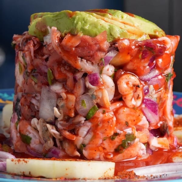 A vibrant Mexican seafood tower with shrimp, octopus, red onion, and avocado, drizzled with red sauce.