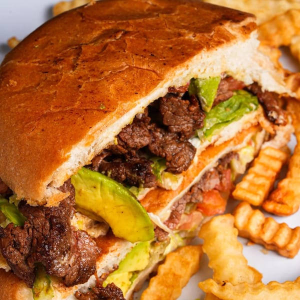 A large Mexican torta sandwich, cut in half, filled with meat, avocado, and lettuce, served with fries.