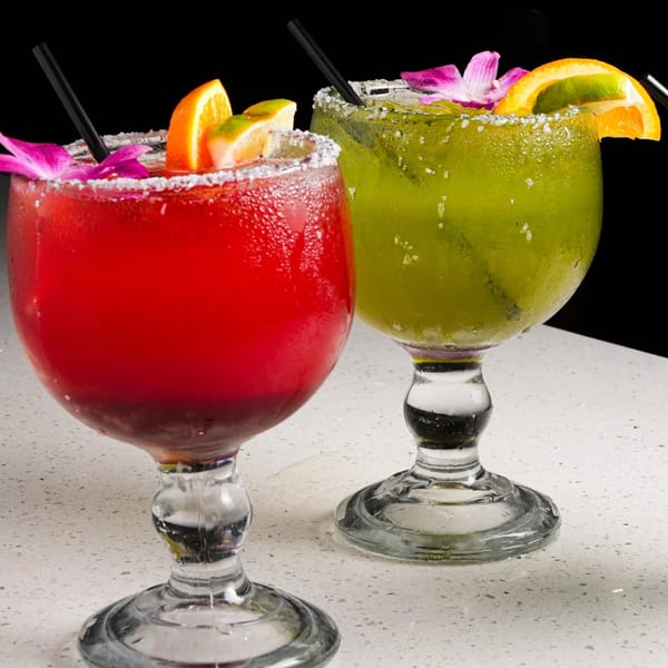 Two El Mezcal margaritas, one red and one green, garnished with fruit and flowers.