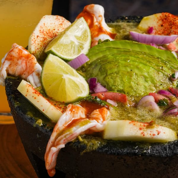 Close-up of a Mexican seafood dish with shrimp, avocado, lime, and red onion in a dark bowl.